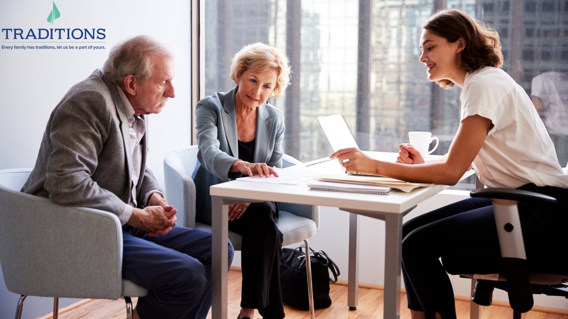 A woman sits across the table and reviews paperwork with an elderly couple with a Traditions logo overlay at top left corner