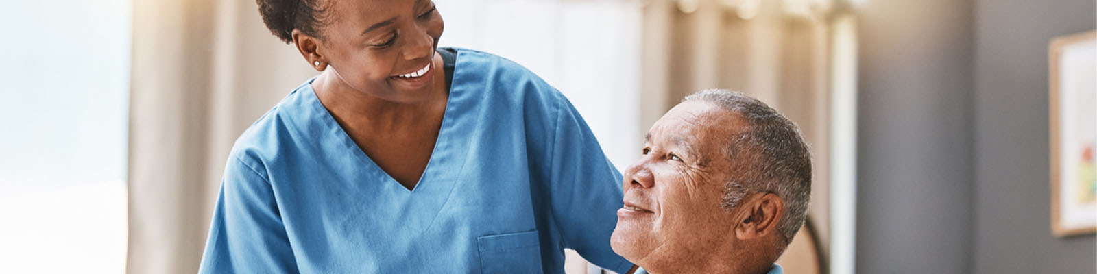 A senior man sitting in a wheelchair looking up at his female nursing assistant with a smile